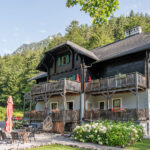 Ein zweistöckiges Haus im Chalet-Stil mit Holzbalkonen, grünen Fensterläden und einer Gartenterrasse vor einer bewaldeten Bergkulisse an einem sonnigen Tag.