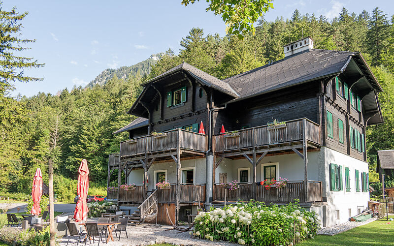 Ein zweistöckiges Haus im Chalet-Stil mit Holzbalkonen, grünen Fensterläden und einer Gartenterrasse vor einer bewaldeten Bergkulisse an einem sonnigen Tag.