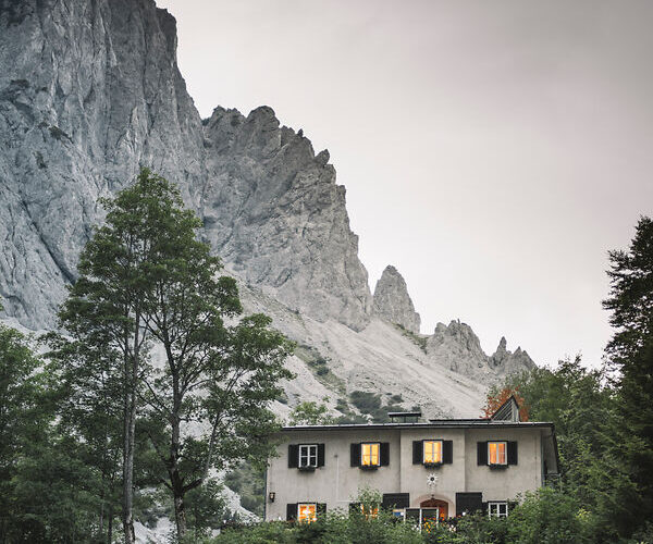 Ein weißes Haus mit beleuchteten Fenstern steht zwischen Bäumen am Fuße eines felsigen Berges unter einem bewölkten Himmel.