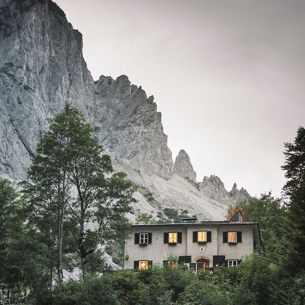 Ein weißes Haus mit beleuchteten Fenstern steht zwischen Bäumen am Fuße eines felsigen Berges unter einem bewölkten Himmel.