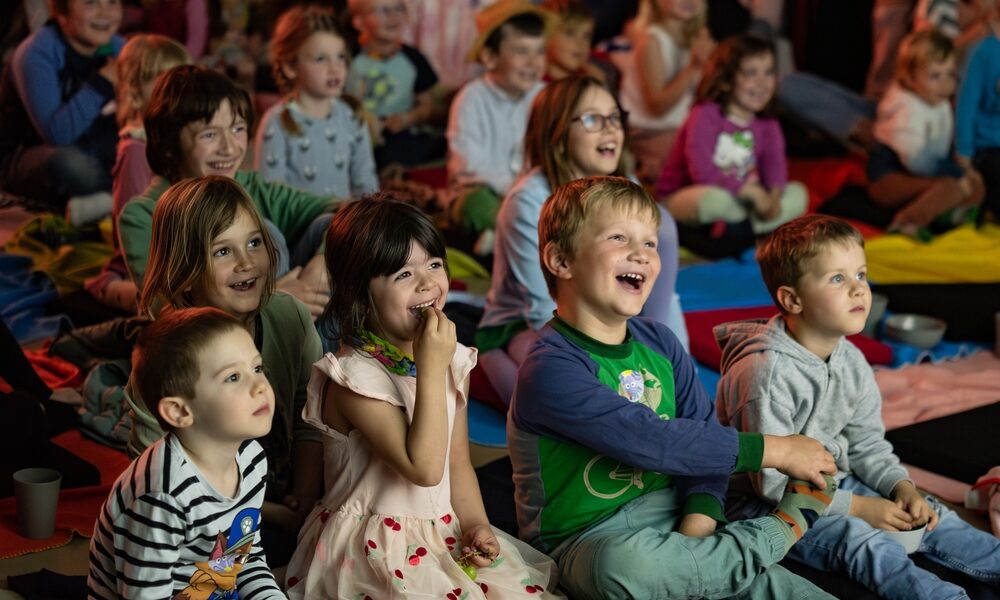 Eine Gruppe kleiner Kinder sitzt eng beieinander auf Matten, lächelt und lacht, während sie etwas aus dem Off beobachten; im Hintergrund sitzen weitere Kinder.