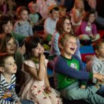 Eine Gruppe kleiner Kinder sitzt eng beieinander auf Matten, lächelt und lacht, während sie etwas aus dem Off beobachten; im Hintergrund sitzen weitere Kinder.