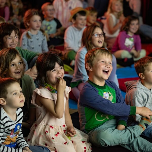 Eine Gruppe kleiner Kinder sitzt eng beieinander auf Matten, lächelt und lacht, während sie etwas aus dem Off beobachten; im Hintergrund sitzen weitere Kinder.