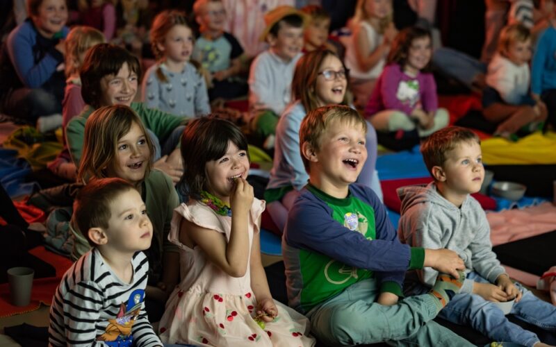 Eine Gruppe kleiner Kinder sitzt eng beieinander auf Matten, lächelt und lacht, während sie etwas aus dem Off beobachten; im Hintergrund sitzen weitere Kinder.