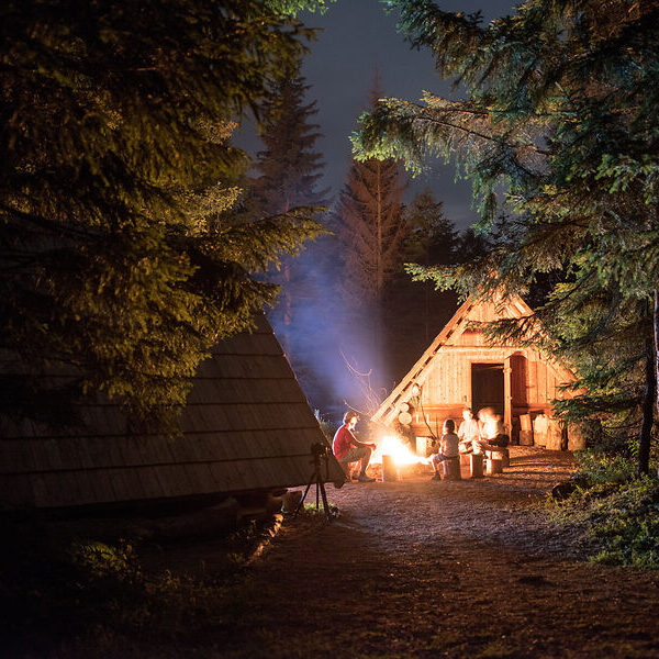 Eine Gruppe von Menschen sitzt nachts um ein Lagerfeuer in der Nähe von Holzhütten in einem Waldgebiet, wobei die Szene von Bäumen umgeben ist.