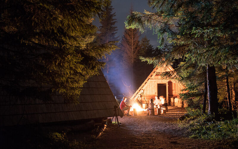 Eine Gruppe von Menschen sitzt nachts um ein Lagerfeuer in der Nähe von Holzhütten in einem Waldgebiet, wobei die Szene von Bäumen umgeben ist.