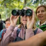 Eine Gruppe von Menschen im Freien, mit einem Kind, das durch ein Fernglas schaut, und einem Erwachsenen, der daneben steht, umgeben von Grünflächen.