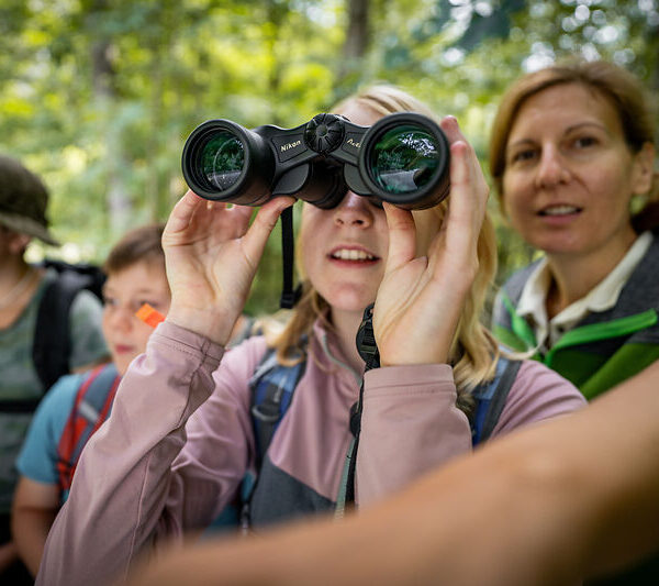Eine Gruppe von Menschen im Freien, mit einem Kind, das durch ein Fernglas schaut, und einem Erwachsenen, der daneben steht, umgeben von Grünflächen.