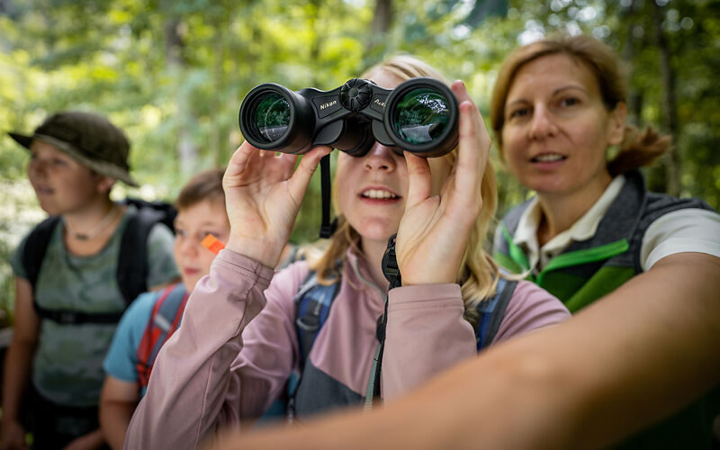 Eine Gruppe von Menschen im Freien, mit einem Kind, das durch ein Fernglas schaut, und einem Erwachsenen, der daneben steht, umgeben von Grünflächen.