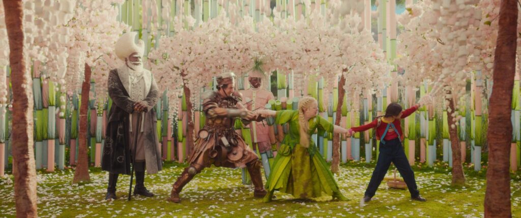 Fünf Personen in aufwendigen, fantasievollen Kostümen stehen in einem Garten mit rosa blühenden Bäumen und hohen, gestreiften Säulen im Hintergrund. Zwei von ihnen zerren am Arm eines Kindes.