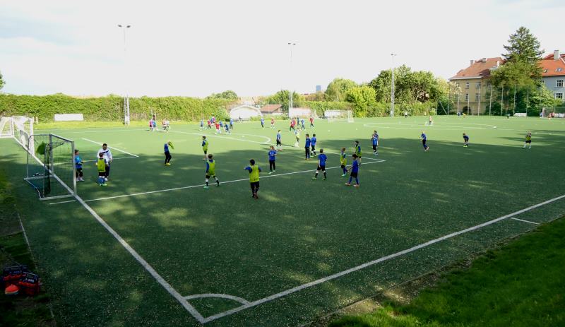 Die Jugendfußballer trainieren auf einem großen grünen Kunstrasenplatz bei Tageslicht, wobei mehrere Gruppen über das Spielfeld verteilt sind und verschiedene Übungen durchführen.