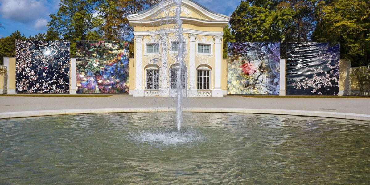 Ein Wasserbrunnen vor einem gelben klassizistischen Gebäude, mit großen abstrakten Kunsttafeln auf beiden Seiten, umgeben von Bäumen unter einem blauen Himmel.