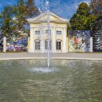 Ein Wasserbrunnen vor einem gelben klassizistischen Gebäude, mit großen abstrakten Kunsttafeln auf beiden Seiten, umgeben von Bäumen unter einem blauen Himmel.