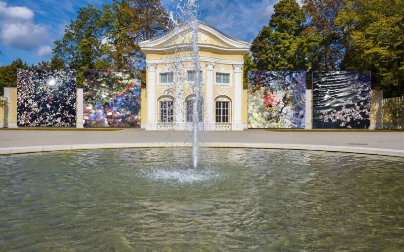 Ein Wasserbrunnen vor einem gelben klassizistischen Gebäude, mit großen abstrakten Kunsttafeln auf beiden Seiten, umgeben von Bäumen unter einem blauen Himmel.