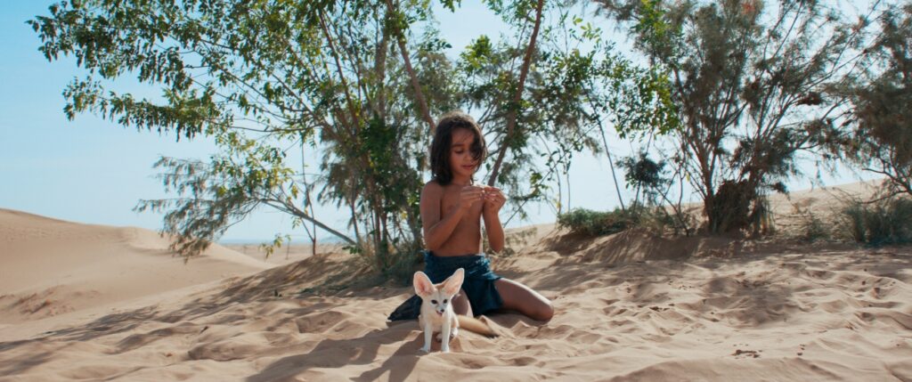 Ein Kind kniet auf sandigem Boden in einer Wüstenlandschaft mit spärlicher Vegetation und hält etwas in den Händen, während ein kleiner Fennec-Fuchs vor ihm sitzt.