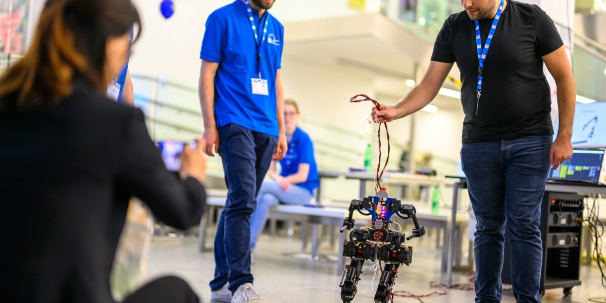 Eine Gruppe von Menschen beobachtet, wie ein Mann in einem schwarzen Hemd einen vierbeinigen Roboter an einem Draht im Haus hält, während eine andere Person ein Foto macht.