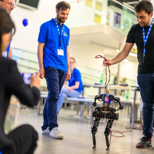 Eine Gruppe von Menschen beobachtet, wie ein Mann in einem schwarzen Hemd einen vierbeinigen Roboter an einem Draht im Haus hält, während eine andere Person ein Foto macht.