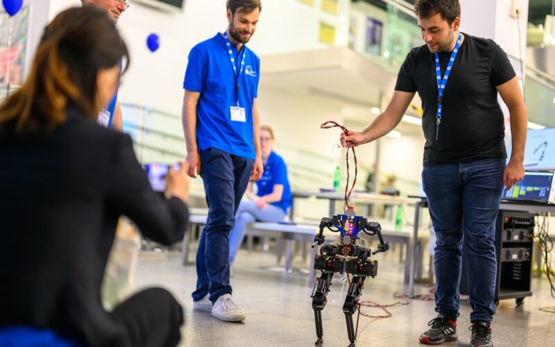 Eine Gruppe von Menschen beobachtet, wie ein Mann in einem schwarzen Hemd einen vierbeinigen Roboter an einem Draht im Haus hält, während eine andere Person ein Foto macht.