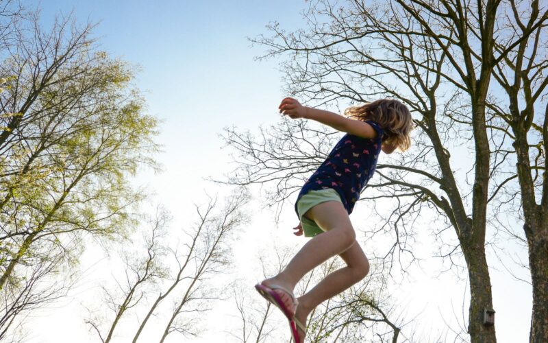 Ein Kind in Shorts und T-Shirt ist mitten im Sprung im Freien, mit Bäumen und einem klaren Himmel im Hintergrund.