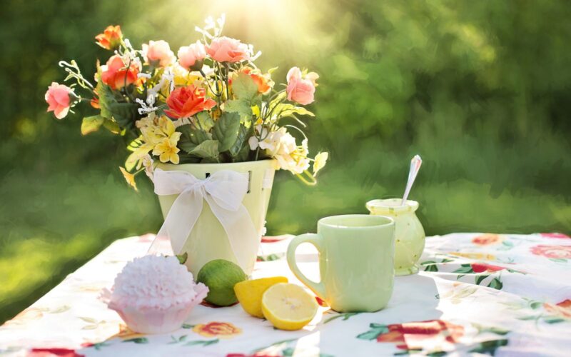 Ein Tisch im Freien mit einer Blumentischdecke, einem Blumenstrauß in einer Vase, einem grünen Becher, einem Glas mit einem Löffel, einem Muffin und einer in Scheiben geschnittenen Zitrone, im Sonnenlicht.