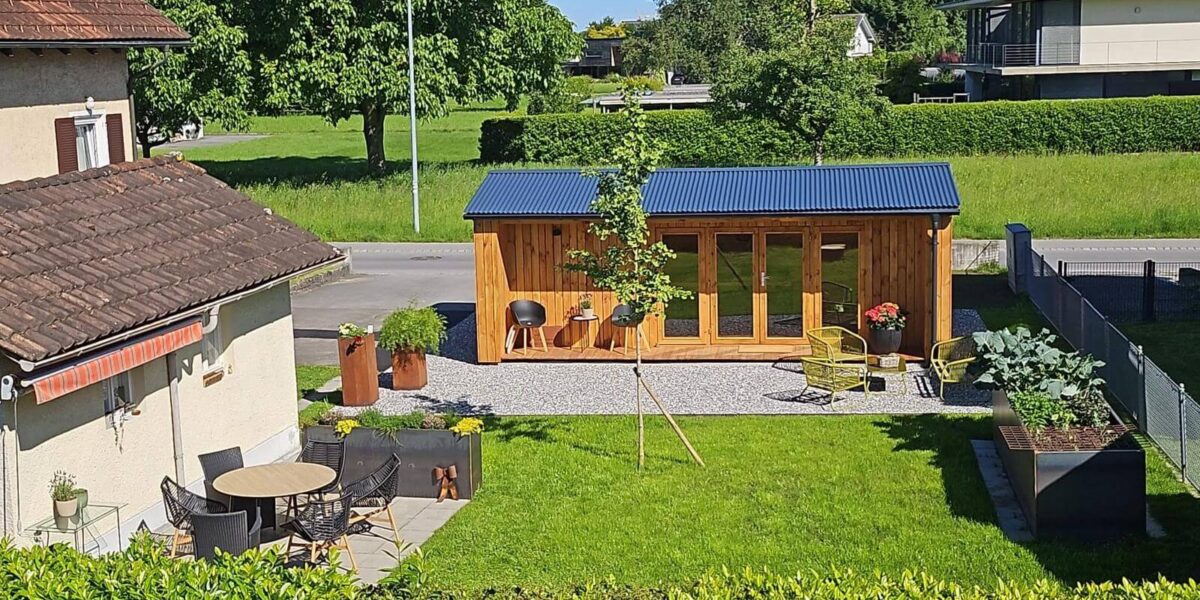 In einem gepflegten Garten neben einem Wohnhaus steht ein moderner Gartenunterstand aus Holz mit blauem Dach, umgeben von Sitzgelegenheiten im Freien, Topfpflanzen und einer kleinen Terrasse.