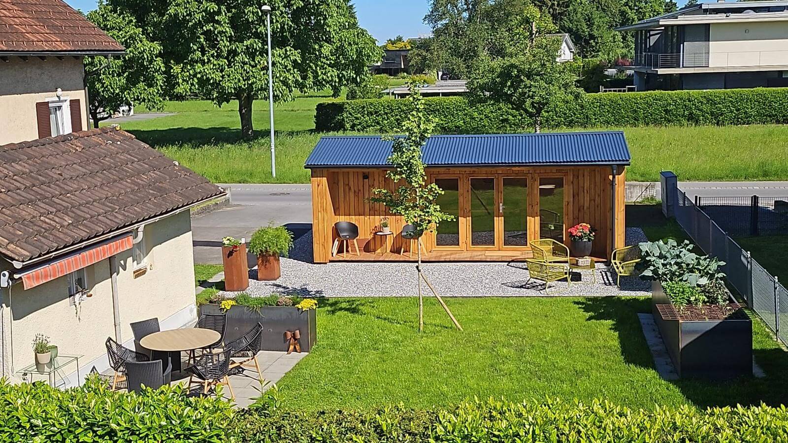 In einem gepflegten Garten neben einem Wohnhaus steht ein moderner Gartenunterstand aus Holz mit blauem Dach, umgeben von Sitzgelegenheiten im Freien, Topfpflanzen und einer kleinen Terrasse.
