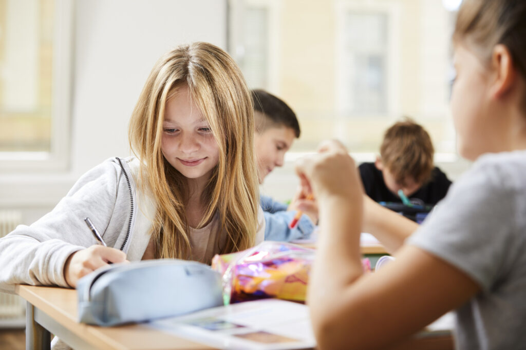 Ein Mädchen schreibt an ihrem Schreibtisch in einem Klassenzimmer, während andere Schüler neben ihr arbeiten. Auf den Schreibtischen liegen Schulmaterialien und Papiere.