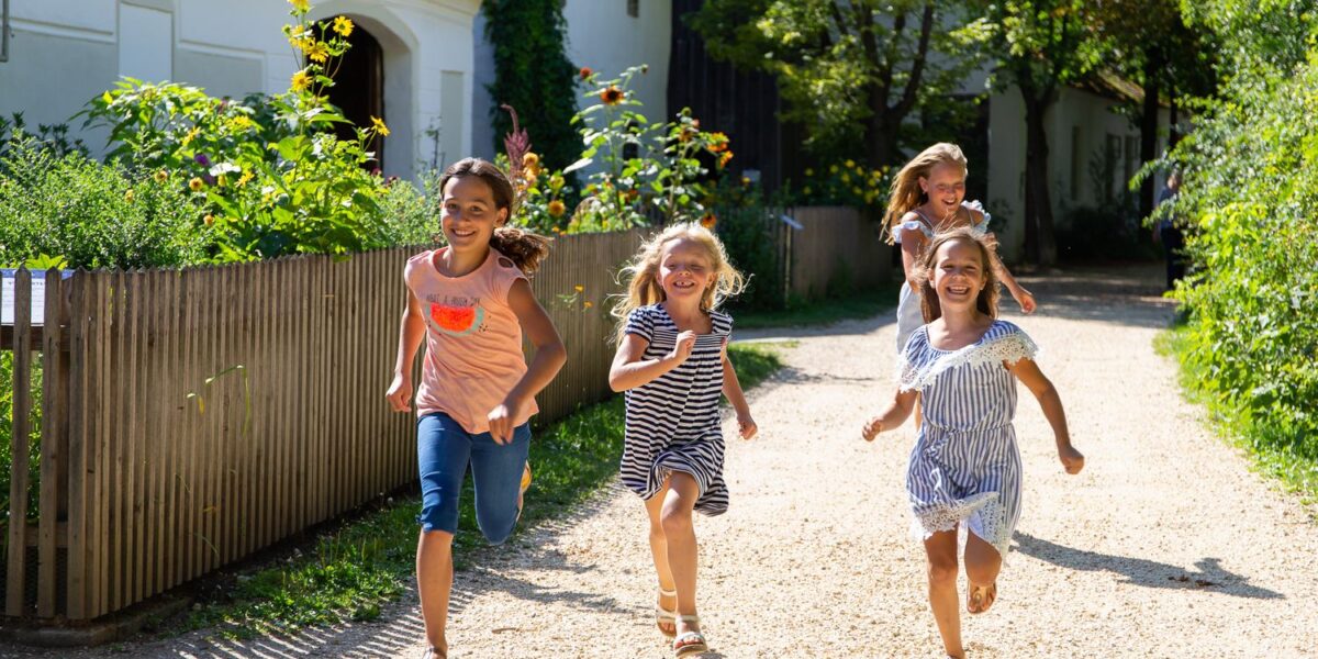 Vier junge Mädchen laufen lächelnd auf einem sonnendurchfluteten Schotterweg neben einem Haus und einem Garten, umgeben von viel Grün.
