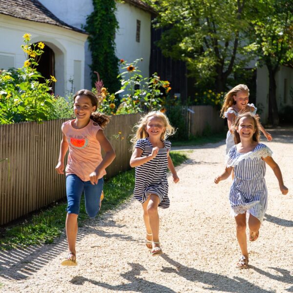 Vier junge Mädchen laufen lächelnd auf einem sonnendurchfluteten Schotterweg neben einem Haus und einem Garten, umgeben von viel Grün.