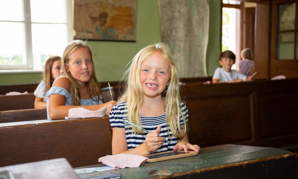 Ein kleines Mädchen lächelt und hält eine Schiefertafel in der Hand, während sie mit anderen Kindern in einem Klassenzimmer an Holzpult sitzt.