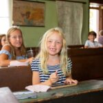 Ein kleines Mädchen lächelt und hält eine Schiefertafel in der Hand, während sie mit anderen Kindern in einem Klassenzimmer an Holzpult sitzt.