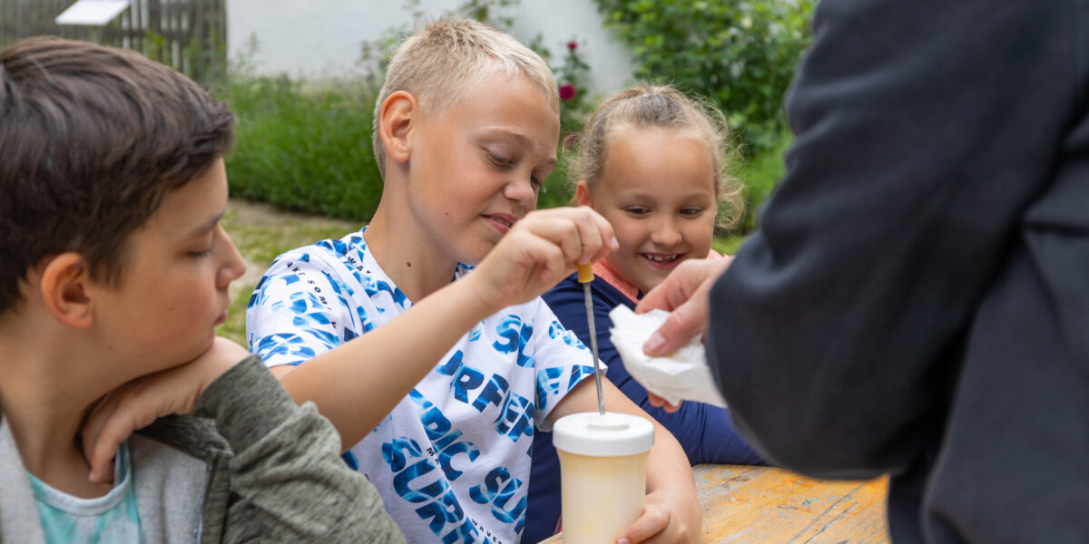 Drei Kinder sitzen an einem Tisch im Freien; eines rührt in einem Getränk um, während ein Erwachsener ihnen etwas reicht. Auf dem Tisch stehen Teller und Tassen; im Hintergrund ist Grün zu sehen.