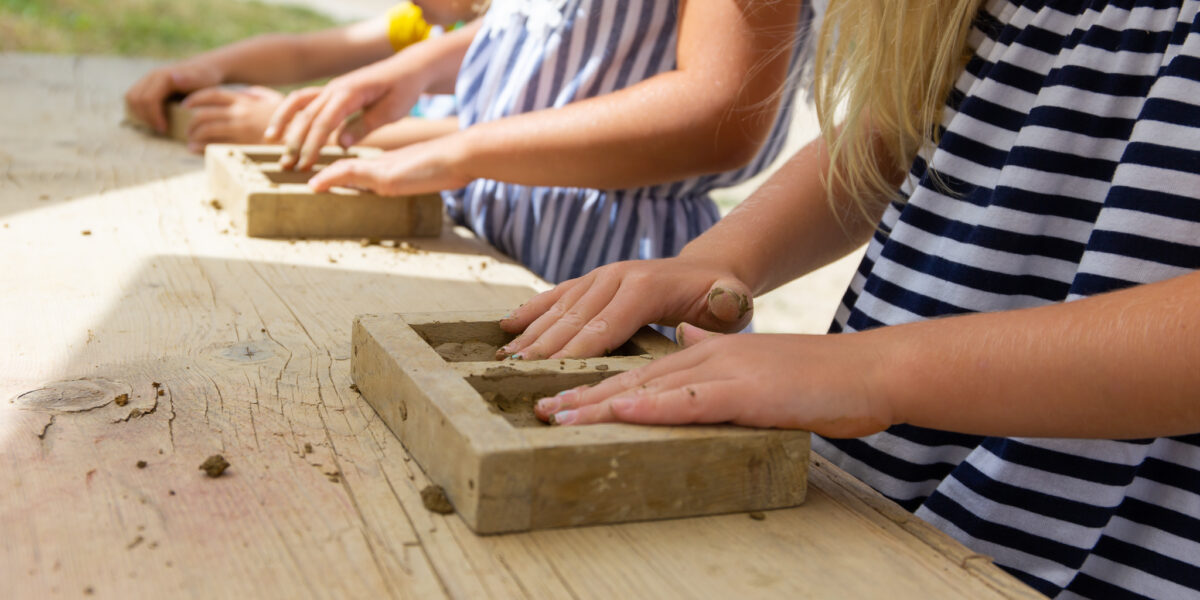 Kinder in gestreiften und gemusterten Kleidern formen mit Hilfe von Holzrahmen Ton auf einem Holztisch im Freien.