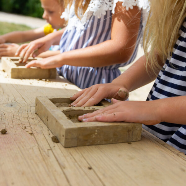 Kinder in gestreiften und gemusterten Kleidern formen mit Hilfe von Holzrahmen Ton auf einem Holztisch im Freien.