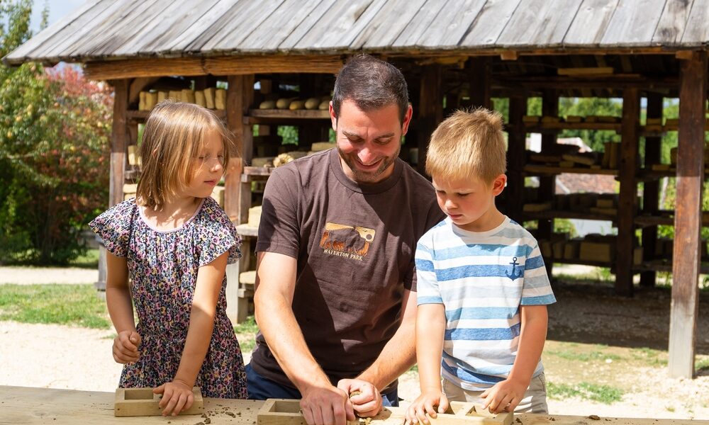 Ein Erwachsener und zwei Kinder arbeiten im Freien an einem Tisch mit Holzrahmen, im Hintergrund sind ein Holzunterstand und Grünflächen zu sehen.