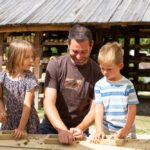 Ein Erwachsener und zwei Kinder arbeiten im Freien an einem Tisch mit Holzrahmen, im Hintergrund sind ein Holzunterstand und Grünflächen zu sehen.