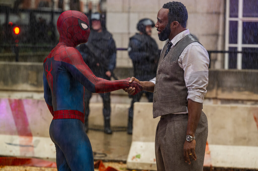 Spider-Man und ein Mann in Weste und Krawatte geben sich draußen im Regen die Hand, im Hintergrund sind Polizisten und Absperrungen zu sehen.