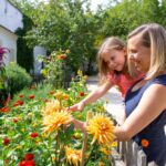 Eine Frau hält ein lächelndes kleines Mädchen im Arm, während die beiden an einem sonnigen Tag in einem farbenfrohen Garten Blumen betrachten und anfassen.