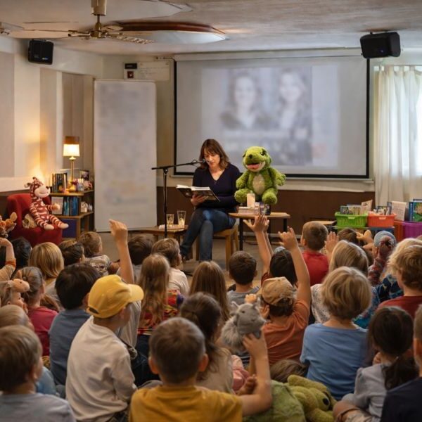 Eine Frau liest einer Gruppe von Kindern vor, die auf dem Boden sitzen, viele von ihnen mit Stofftieren in der Hand, mit einer grünen Puppe neben ihr und einer unscharfen Projektion im Hintergrund.