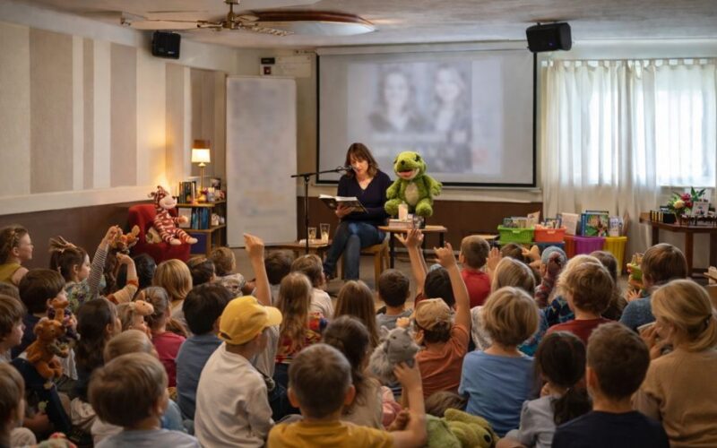 Eine Frau liest einer Gruppe von Kindern vor, die auf dem Boden sitzen, viele von ihnen mit Stofftieren in der Hand, mit einer grünen Puppe neben ihr und einer unscharfen Projektion im Hintergrund.