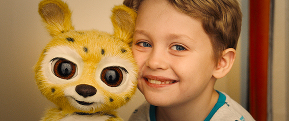 Ein kleiner Junge lächelt neben einem großen, gelben Plüschtier mit großen Augen und Flecken.