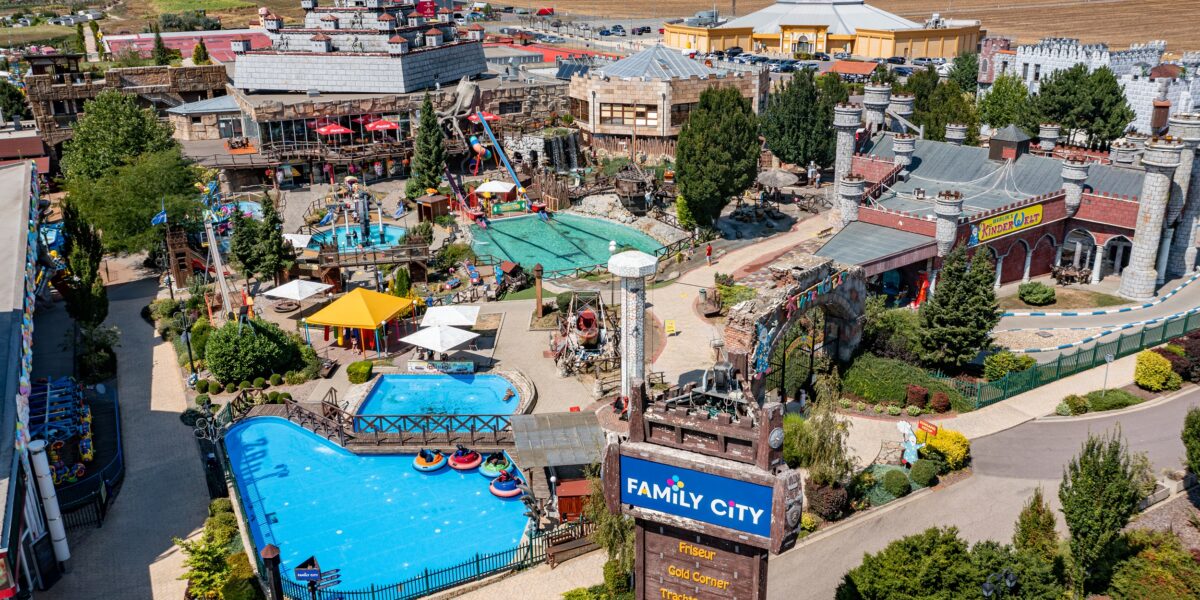 Luftaufnahme eines Vergnügungsparks mit Schwimmbecken, Fahrgeschäften, landschaftlich gestalteten Wegen und Gebäuden. In der Nähe des Eingangs ist ein Schild mit der Aufschrift „Family City“ zu sehen. Der Park ist von Feldern und Gebäuden umgeben.