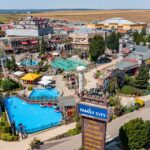 Luftaufnahme eines Vergnügungsparks mit Schwimmbecken, Fahrgeschäften, landschaftlich gestalteten Wegen und Gebäuden. In der Nähe des Eingangs ist ein Schild mit der Aufschrift „Family City“ zu sehen. Der Park ist von Feldern und Gebäuden umgeben.