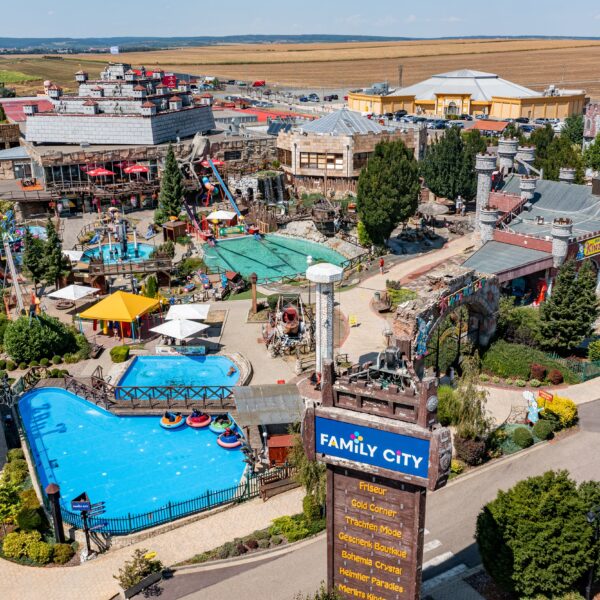 Luftaufnahme eines Vergnügungsparks mit Schwimmbecken, Fahrgeschäften, landschaftlich gestalteten Wegen und Gebäuden. In der Nähe des Eingangs ist ein Schild mit der Aufschrift „Family City“ zu sehen. Der Park ist von Feldern und Gebäuden umgeben.