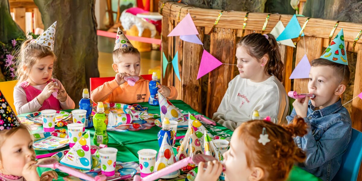 Kinder mit Partyhüten sitzen bei einer Geburtstagsfeier um einen geschmückten Tisch herum, blasen in Trillerpfeifen und sind von bunten Tellern, Bechern und Girlanden umgeben.
