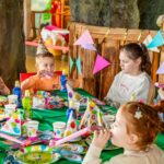 Kinder mit Partyhüten sitzen bei einer Geburtstagsfeier um einen geschmückten Tisch herum, blasen in Trillerpfeifen und sind von bunten Tellern, Bechern und Girlanden umgeben.