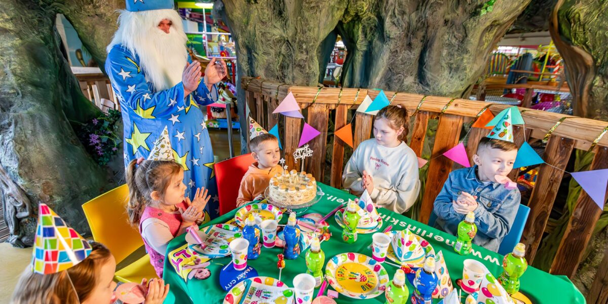 Ein Zauberer unterhält sechs Kinder auf einer Geburtstagsparty mit Dekorationen, Partyhüten und einer Torte auf einem bunten Tisch in einem Raum, der im Baum-Stil gestaltet ist.