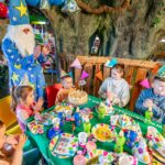 Ein Zauberer unterhält sechs Kinder auf einer Geburtstagsparty mit Dekorationen, Partyhüten und einer Torte auf einem bunten Tisch in einem Raum, der im Baum-Stil gestaltet ist.