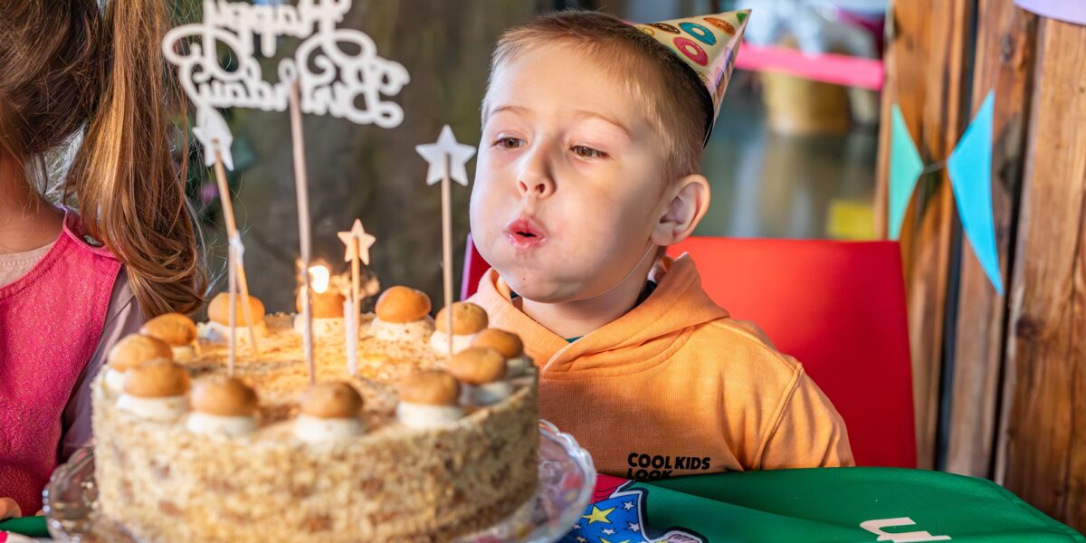 Ein kleiner Junge mit einem Partyhut bläst bei einer Feier die Kerzen auf einer geschmückten Geburtstagstorte aus.