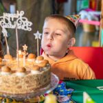Ein kleiner Junge mit einem Partyhut bläst bei einer Feier die Kerzen auf einer geschmückten Geburtstagstorte aus.